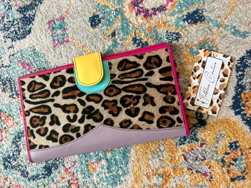 A leopard print wallet with a lavender side panel, hot pink trim, a yellow and turquoise snap closure, and a branded tag reading 'Fiction Custom' placed on a patterned rug.